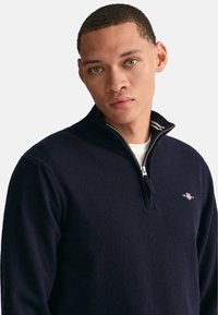Marineblauwe gebreide pullover met een halve rits bij de kraag, voorzien van een klein logo op de borst en contrasterende lichte strepen op de kraag. Zachte textuur.