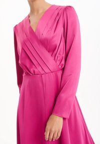 Robe en satin fuchsia à manches longues, avec un décolleté en V croisé et des détails plissés sur le corsage. Texture lisse.