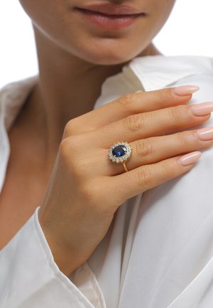 Hand mit gepflegten Nägeln, die einen Goldring mit großem ovalem blauem Saphir trägt, umgeben von kleinen Diamanten, auf der Schulter, weiße Bluse sichtbar.