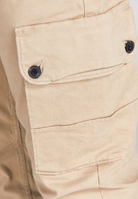 Poche sur un pantalon cargo en tissu beige avec deux boutons noirs assurant la fermeture à rabat sur le côté de la jambe.