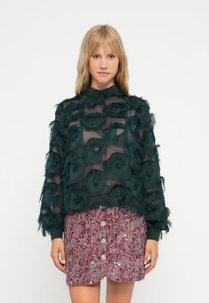Top verde trasparente con applicazioni floreali testurizzate, abbinato a una gonna rosa con paillettes caratterizzata da tre grandi bottoni sul davanti.