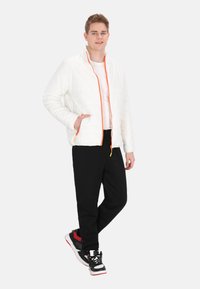 Jachetă albă matlasată cu fermoar și accente portocalii, purtată peste un top alb, asortată cu pantaloni negri și adidași multicolori.