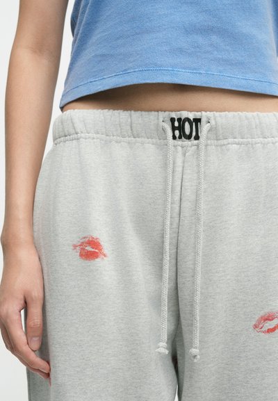 Calças de treino cinzentas com estampas de lábios vermelhos e uma cintura elástica com a palavra "HOT" impressa a preto. Também apresentam cordões de ajuste.