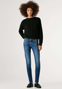 Mustast kootud lahtise lõikega kampsun, mida on kombineeritud siniste kitsaste teksade ja mustade paeltega kingadega. Minimalistlik disain, ilma mustrite või nähtavate detailideta.