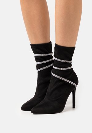 Botines de tacón alto de ante negro con puntera afilada, adornados con múltiples cintas de strass plateadas envueltas alrededor de los tobillos.