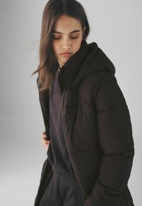 Doudoune noire avec capuche, fermeture éclair à l'avant et texture matelassée. Portée sur un haut foncé, présentant une coupe ajustée et un tissu doux.