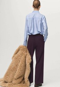 Personne avec les cheveux en chignon portant une chemise bleu clair et un pantalon noir, tenant un grand manteau en fausse fourrure beige qui tombe jusqu'au sol.