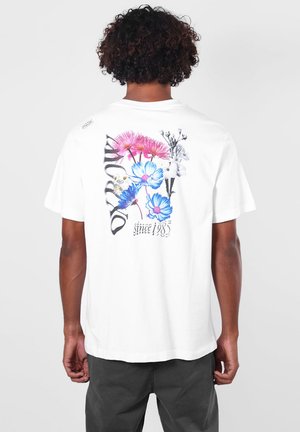 Personne aux cheveux bouclés portant un T-shirt blanc avec des motifs colorés de fleurs roses et bleues et le texte "since 1985" dans le dos.