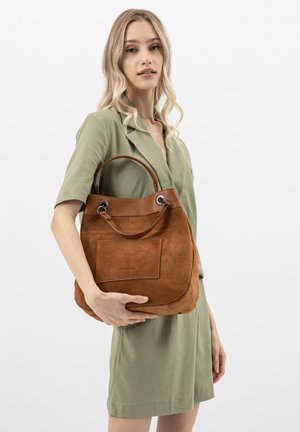 Młoda kobieta w jasnooliwkowej sukience trzyma dużą torbę z brązowego zamszu z skórzanymi wykończeniami na białym tle.