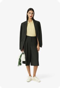 Blazer oversize a quadri verde scuro, polo shirt giallo chiaro, pantaloni corti a pieghe, sneakers bianchi e borsa bianca con una sciarpa verde.