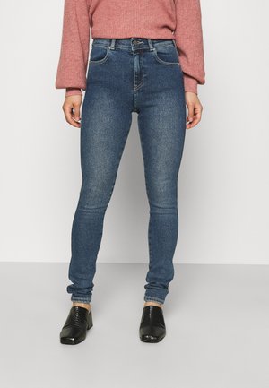 Personne portant un jean bleu ajusté, un pull rose à manches longues et des chaussures noires à talons, debout devant un fond clair uni.