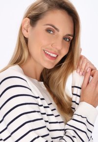 Weißer Cardigan mit navy-blauen horizontalen Streifen und Knopfverschluss, mit einer weichen Textur. Das Model hat lange, lockige blonde Haare und einen neutralen Hintergrund.