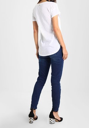 Camiseta blanca de manga corta con dobladillo redondeado, combinada con jeans ajustados azul oscuro y tacones negros con lunares blancos y detalle de lazo en la parte trasera.