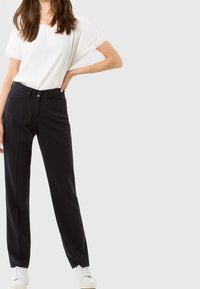 Kobieta ubrana w białą bluzkę z krótkim rękawem, czarne spodnie typu straight-leg i białe trampki, stoi z jedną ręką na biodrze.