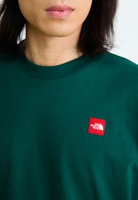Hanorac verde din bumbac, cu guler rotund și manșete ribbrate. Prezintă o etichetă mică roșie și albă pe piept. Textură netedă.
