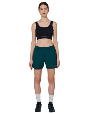 Kobieta stojąca, ubrana w czarny sportowy biustonosz, ciemnozielone krótkie spodenki sportowe, białe skarpetki i czarne buty do biegania na białym tle.