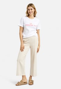 White cotton t-shirt with "SUNSHINE CLUB" printed in pink, paired with beige wide-leg trousers and tan slide sandals with ruffle accents.