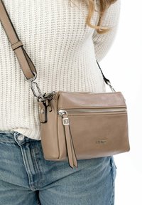 Bolso cruzado de cuero beige con herrajes plateados, cierre de cremallera y un bolsillo delantero. Correa ajustable y detalle del logo sutilmente visible.