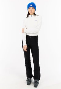 Hvid langærmet zip-up trøje med et logo, sorte skibukser med lommer, blå strikket hue og grå skistøvler. Enkel, funktionel vinterbeklædning.