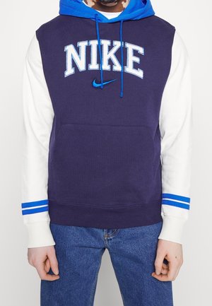 Osoba ubrana w granatowo-białą bluzę Nike z kapturem w kolorze niebieskim i mankietami w niebieskie paski, w połączeniu z niebieskimi dżinsami.