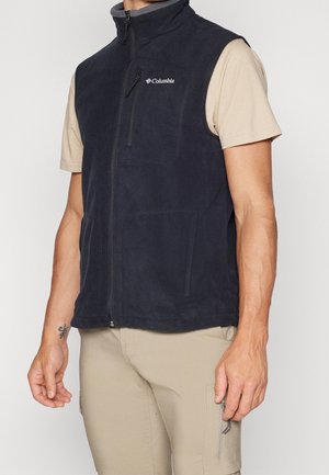 Mann trägt eine schwarze Columbia-Fleeceweste mit Reißverschluss über einem beige-farbenen kurzärmligen Hemd und khakifarbenen Hosen, steht mit entspannt an den Seiten hängenden Händen.