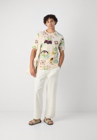 Short-sleeve beige t-shirt with colorful floral and eye graphics, paired with white wide-leg pants and beige sandals, carried with a canvas bag.