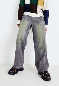 Jeans anchos grises con un acabado desgastado, que presentan un corte holgado y detalles de costura sutiles, combinados con zapatillas texturizadas en negro y naranja.