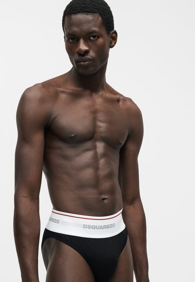 Μυώδης μαύρος άνδρας που φοράει μαύρο boxer με λευκή μέση με την επιγραφή «DSQUARED2», στέκεται μπροστά σε μια απλή λευκή επιφάνεια.