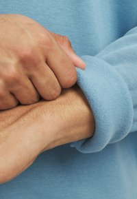 Na hlačah je prikazan manšeta svetlo modrega flis jopiča, ki se ponaša z mehkobo in rahlo sproščenim krojem. Vidna roka drži manšeto.