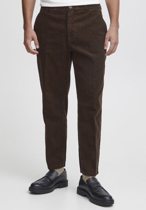 Pantaloni in corduroy marrone con una vestibilità a gamba dritta, dotati di chiusura con bottone e due tasche laterali. Abbinati a scarpe nere slip-on.