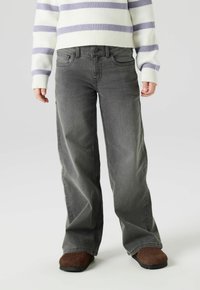 Persoon draagt losse grijze spijkerbroek, bruine zachte slippers en een witte trui met paarse horizontale strepen op een effen achtergrond.