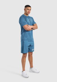 Niebieska koszulka sportowa z krótkimi rękawami, w połączeniu z niebieskimi szortami z białym logo. Całość dopełniają białe trampki.
