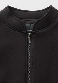 Veste noire à fermeture éclair avec col haut, confectionnée dans un tissu doux. Présente une fermeture éclair argentée et un design minimaliste. Étiquette de la marque à l'intérieur du col.