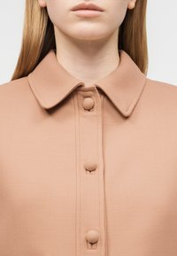 Γυναίκα που φοράει μπεζ πουκάμισο με γιακά και κουμπιά καλυμμένα με ύφασμα, εμφανιζόμενη από τον λαιμό έως το πάνω μέρος του στήθους, σε απλό φόντο.