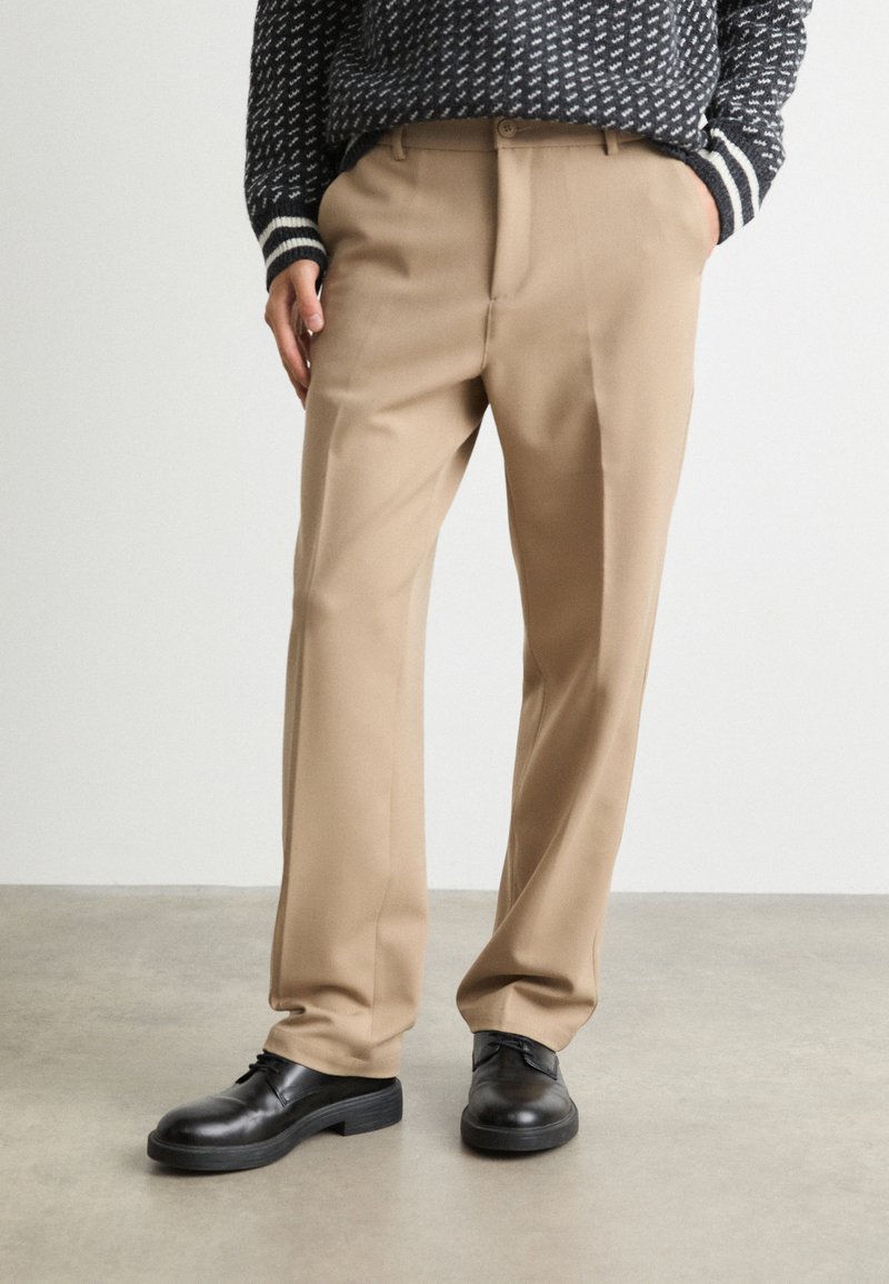 Pantalones beige con ajuste entallado, hechos de una tela suave. Cuentan con una cinturilla clásica y bolsillos laterales, combinados con zapatos negros.