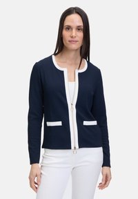 Gilet zippé bleu marine avec des accents blancs, col rond, deux poches avant, tissu texturé et fermoir en métal argenté.