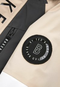 Beżowa i czarna kurtka z gładkiego materiału, z wyraźną okrągłą naszywką z logo i zamkiem błyskawicznym, wykończona czarnym napisem.