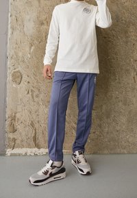 Weißes Langarmhemd mit Logo, blaue Sporthose und graue sowie burgunderfarbene Sneaker mit strukturiertem Obermaterial und sichtbaren Markendetails.