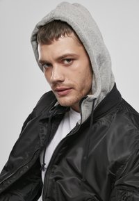 Jachetă bomber neagră cu căptușeală de glugă gri. Jacheta are un închidere cu fermoar și șnururi. Textură netedă și lucioasă.