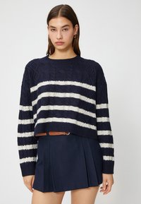 Niebiesko-cremowy sweter z prążkowanym wzorem, skrócony, z teksturą warkocza, z długimi rękawami i okrągłym dekoltem, w zestawieniu z plisowaną niebieską spódnicą.