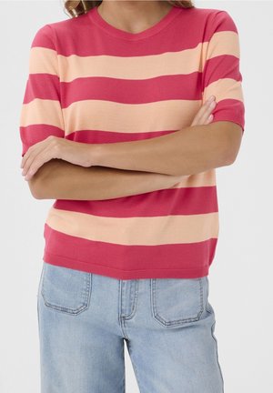 Kobieta ze skrzyżowanymi ramionami, ubrana w krótki sweter z szerokimi, koralowymi i brzoskwiniowymi poziomymi paskami oraz jasnoniebieskie dżinsy.
