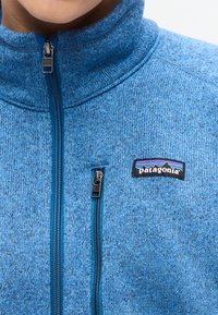 Μπλε πλεκτό φλις μπουφάν Patagonia με πλήρες φερμουάρ μπροστά και τσεπάκι στο στήθος με φερμουάρ, φορεμένο από άτομο από τον λαιμό έως το μέσο του στήθους.
