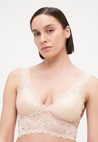 Lace bralette in light nude with floral patterns, V-neck design, wide straps, and a scalloped hem. Soft texture and delicate detail.