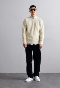 Light beige ribbed sweater, white button-up shirt beneath, black pants, and white sneakers. The person wears sunglasses, standing against a gray background.