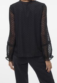 Blouse noire avec manches transparentes à pois, poignets volantés et coupe fluide. Tissu lisse avec des détails texturés sur l'ensemble.