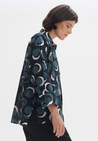 Camicia a maniche lunghe con bottoni, caratterizzata da uno sfondo scuro con grandi motivi circolari in turchese e bianco. Realizzata in un tessuto leggero con una vestibilità morbida.
