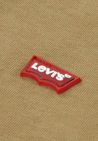 Patch logo Levi's en caoutchouc rouge avec lettrage blanc, cousu sur un fond de tissu beige texturé. Le patch a une forme et un design classiques.