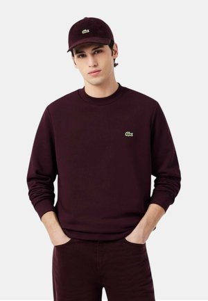 Uomo che indossa una camicia a maniche lunghe bordeaux scuro, pantaloni abbinati e un berretto di velluto a coste, tutti con un piccolo logo verde del coccodrillo sul petto e sul berretto.