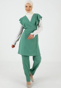 Vihreä V-pääntie mekko, jossa on röyhelösateet, yhdistettynä samanvärisiin housuihin. Materiaali on sileä; mukana on vaaleanvärinen huivi ja beige korkokengät.