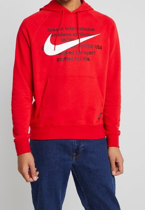 Persoon die een rode Nike hoodie met wit swoosh-logo en zwarte tekst draagt, gecombineerd met blauwe jeans, staand tegen een effen achtergrond.